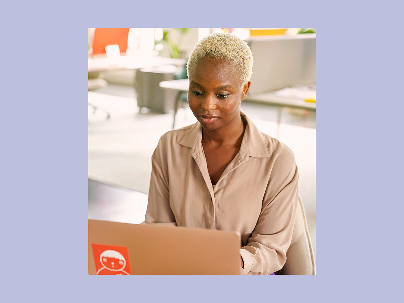 Person with short light hair wearing a beige shirt sitting at a desk using a laptop with a red sticker in a bright office environment