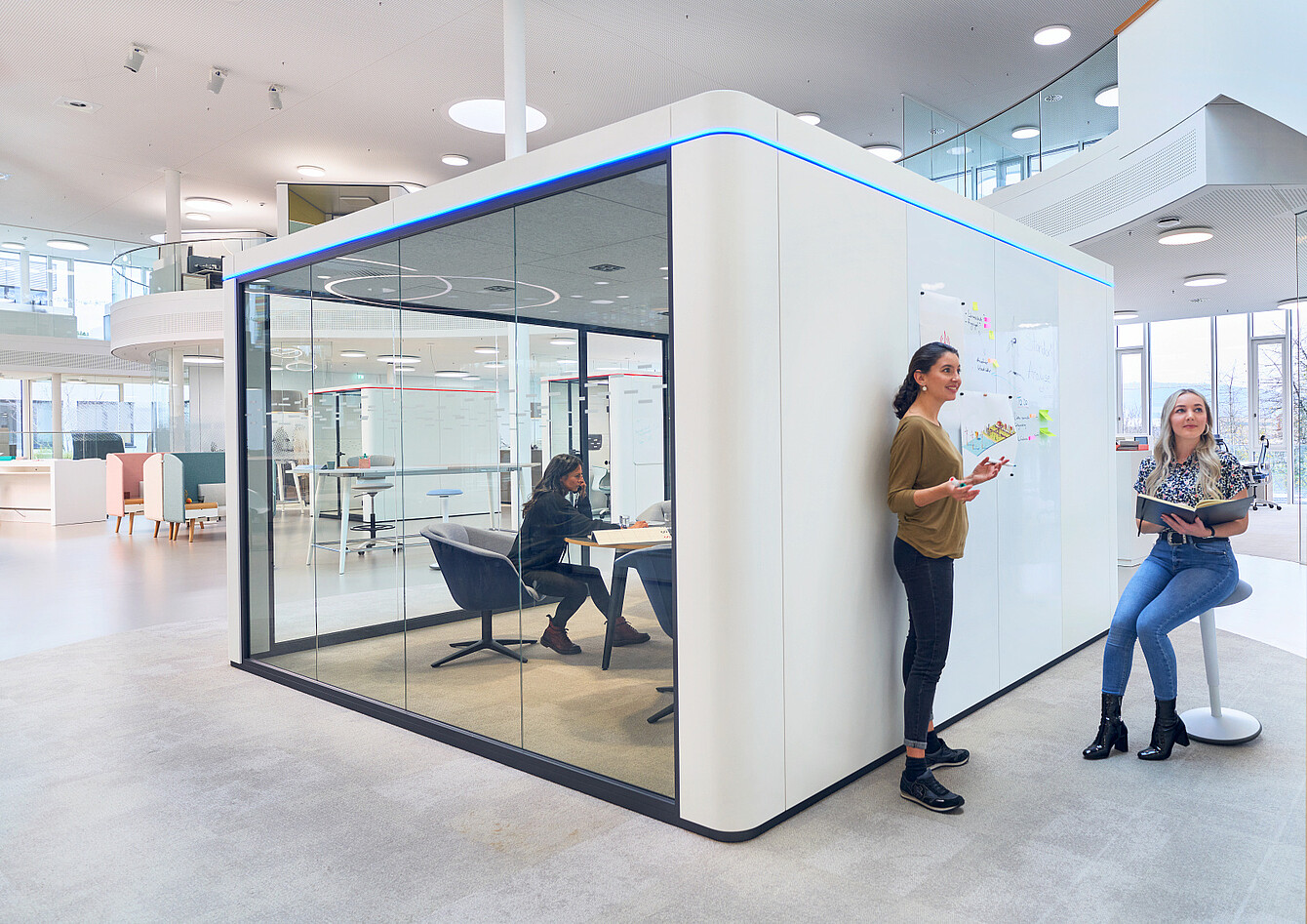 Modern office space with a glass-walled meeting room and two women standing and sitting near a whiteboard wall with notes
