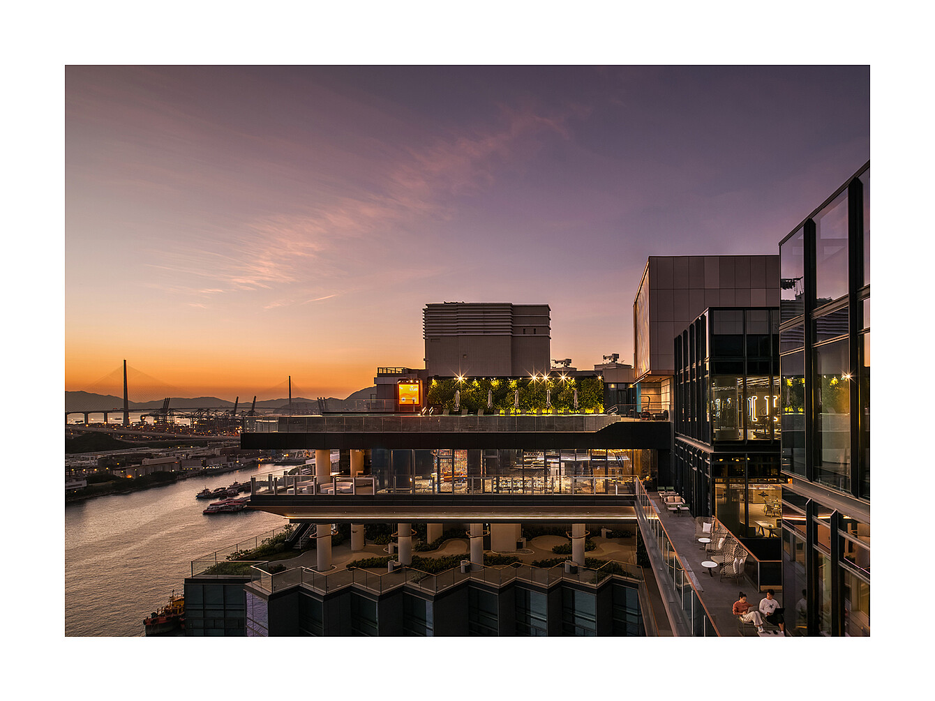 Modern multi-story building with terraces and glass walls overlooking a river at sunset