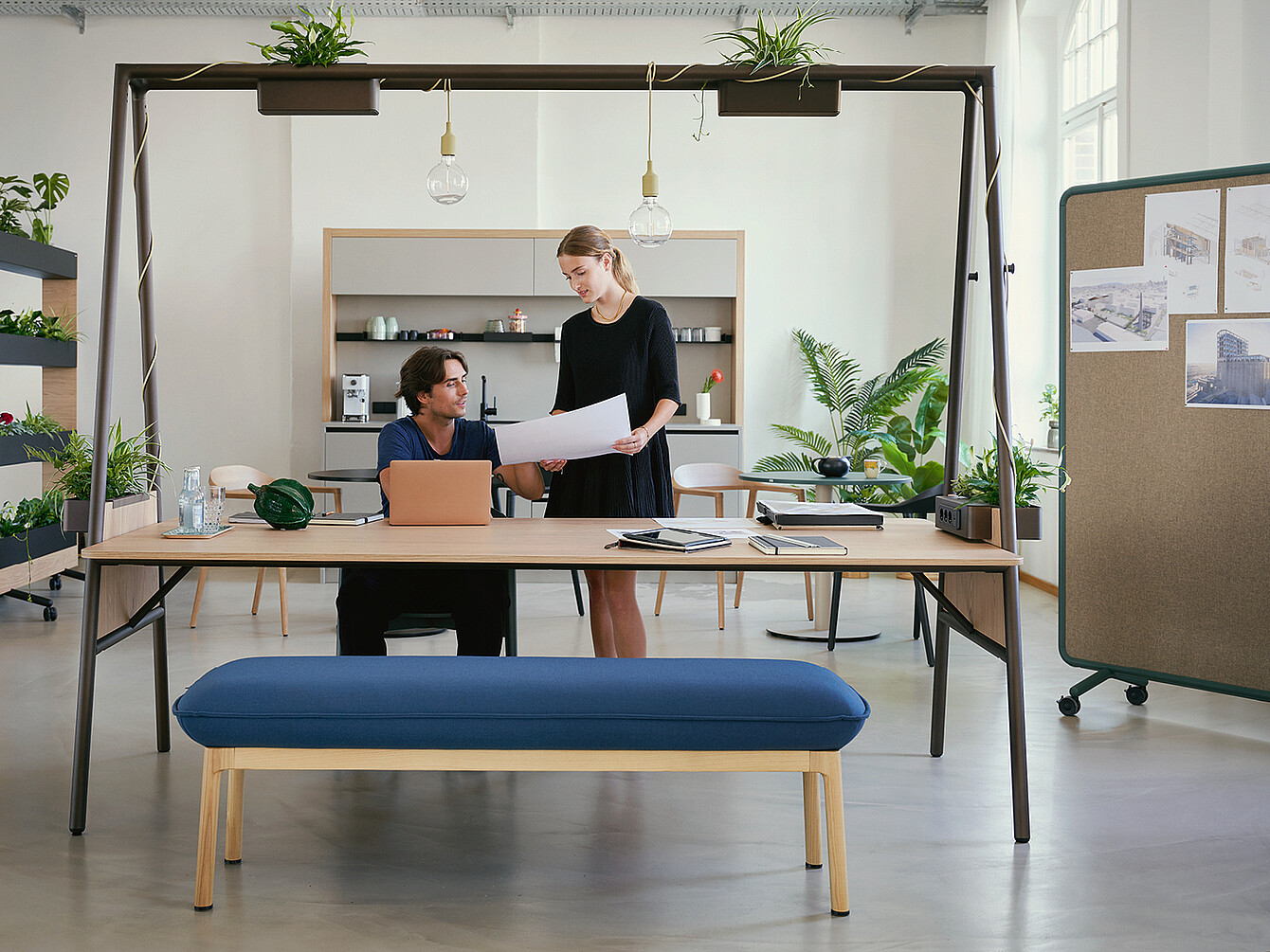 Zwei Personen im modernen Work Café Büro, eine sitzt mit Laptop, die andere steht mit Papier, umgeben von Pflanzen und Designermöbeln