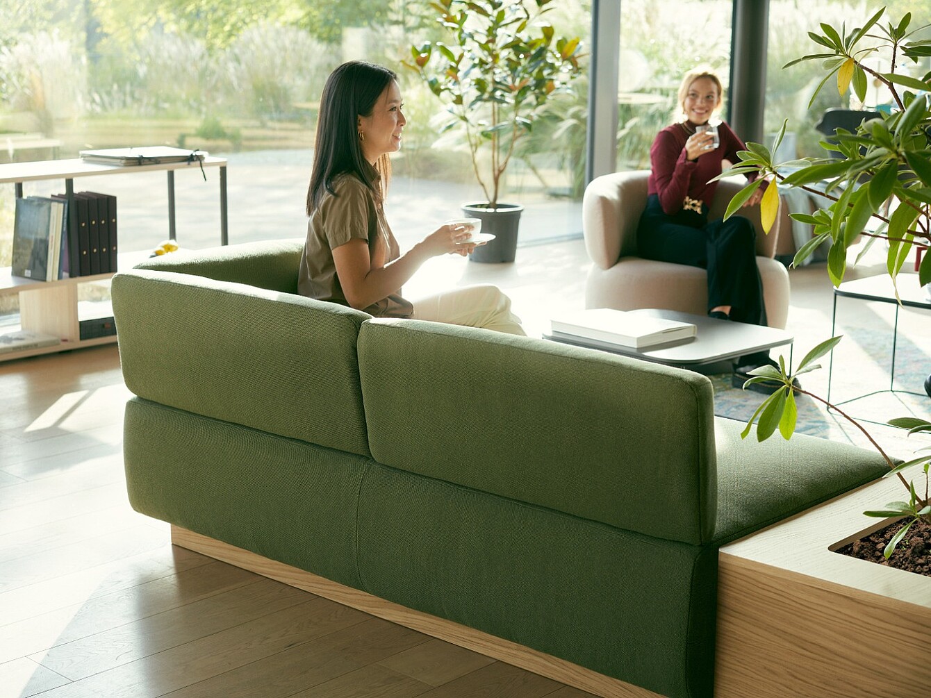 Two women sit on sofas in a bright room drinking from cups