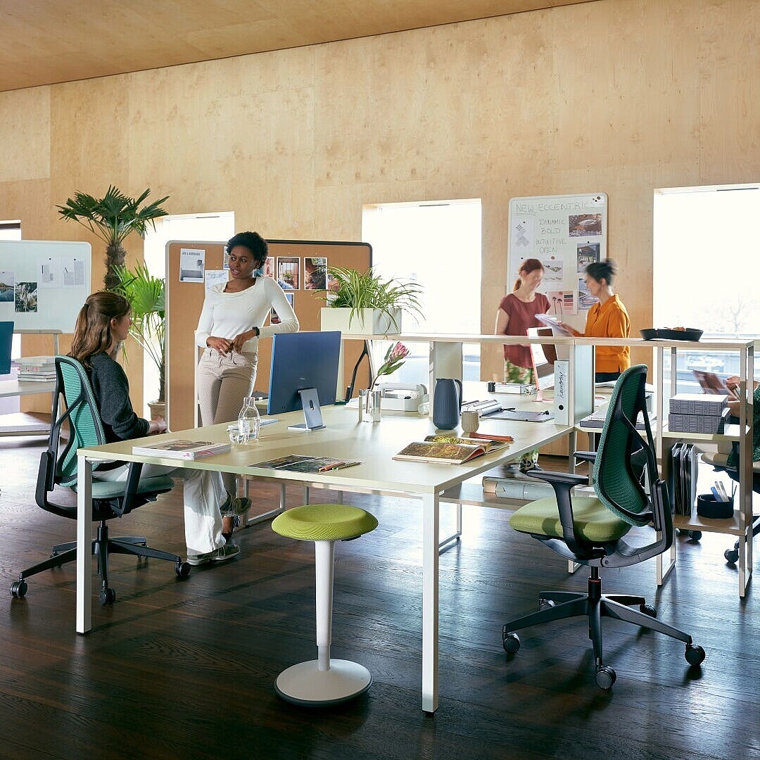 Offenes Büro mit mehreren Personen, die an höhenverstellbaren Tischen arbeiten und sich bewegen, um aktive Pausen und dynamisches Sitzen zu fördern.