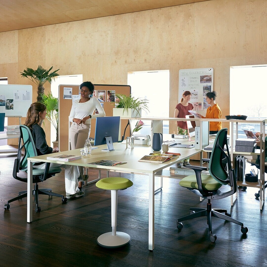 Open-plan office with people working at desks and standing, featuring ergonomic chairs, stools, and plants