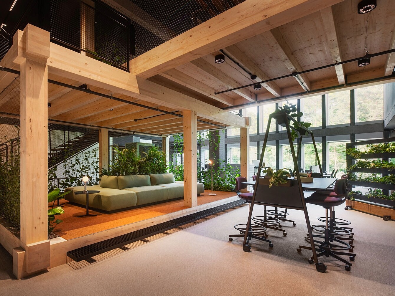 Moderner Arbeitsbereich im Novu Campus mit großem Holzbalkenrahmen, grüner Couch, mehreren Stühlen und Pflanzen vor großen Fenstern.