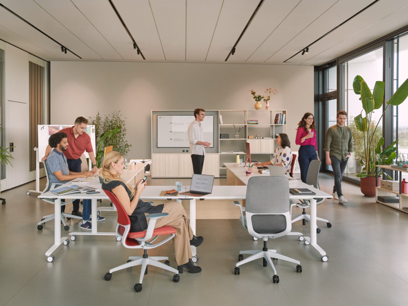 Office scene with several people sitting, standing, and talking at desks in a bright room with large windows and plants