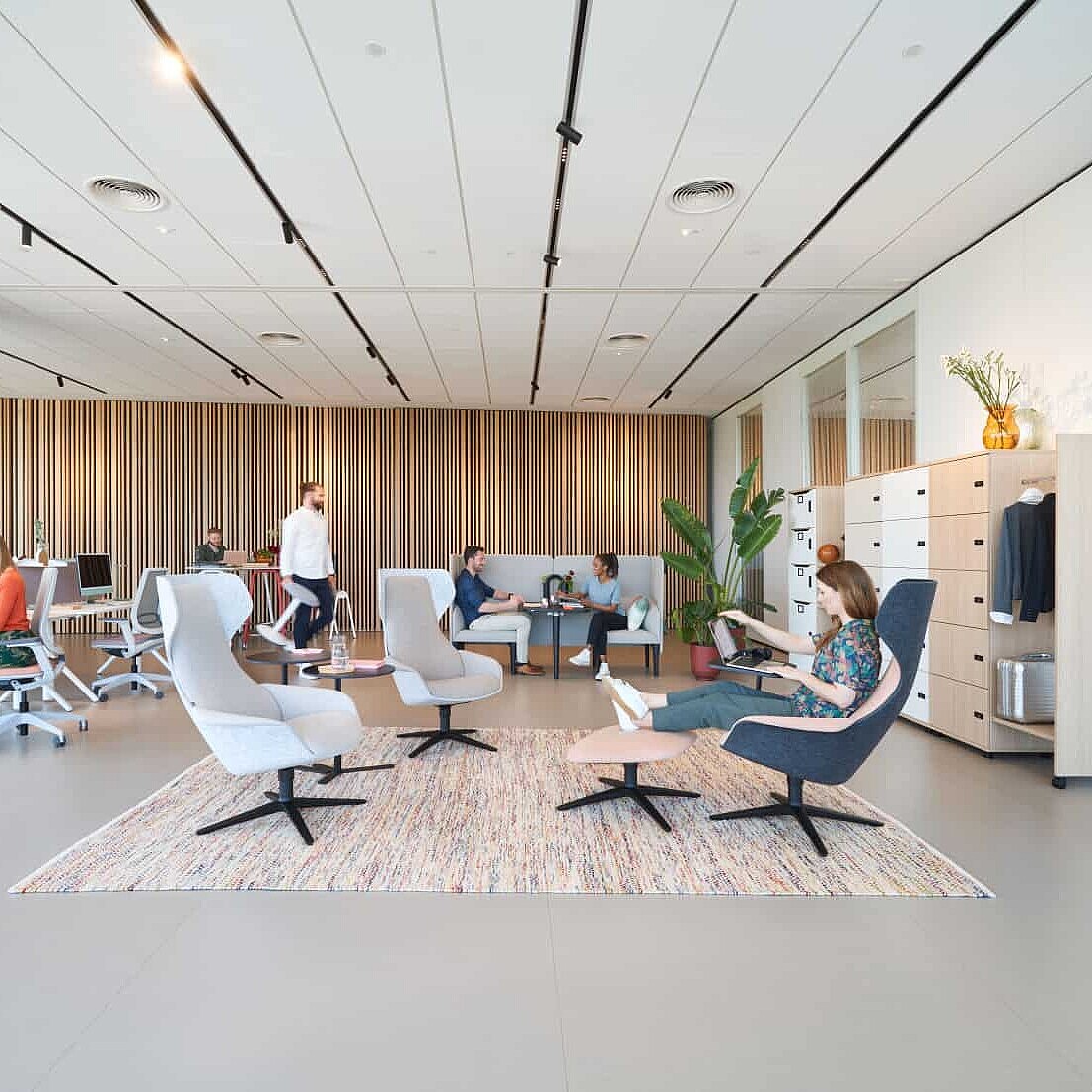 Eine offene Bürofläche mit modernen Möbeln, die eine entspannte Arbeitsumgebung fördern. Mehrere Personen arbeiten in einem großzügigen, lichtdurchfluteten Raum, der mit ergonomischen Stühlen und bequemen Sitzbereichen ausgestattet ist. Pflanzen und Holzelemente verleihen dem Raum eine natürliche Atmosphäre. SEO-Schlüsselwörter: "hybrides Büro", "moderne Arbeitswelt", "flexible Arbeitsumgebungen", "Büro mit Wohlfühlatmosphäre".