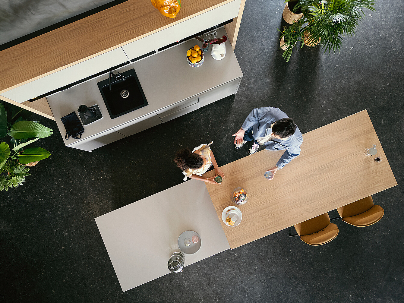 Overhead view of two people conversing at a wooden table in a modern hybrid ductile commons workspace with integrated kitchen area and plants