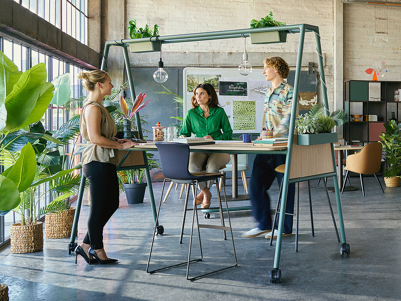 Three colleagues collaborate around a high table with plants on top in a bright office filled with greenery, exemplifying a biophilic Garden Work Café design.