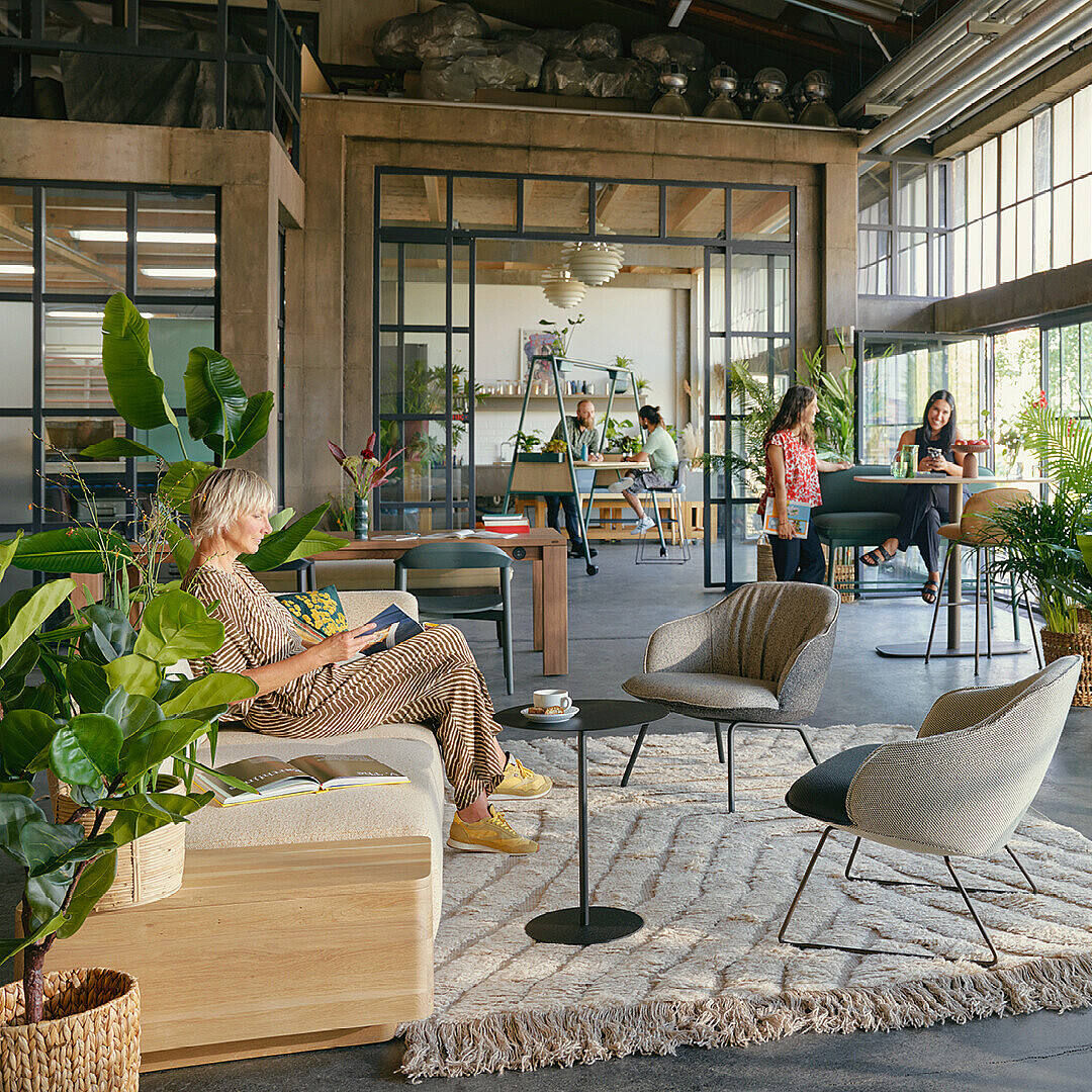 Open office space with large windows, abundant indoor plants, and modern seating creating a Garden Work Café environment inspired by biophilic design