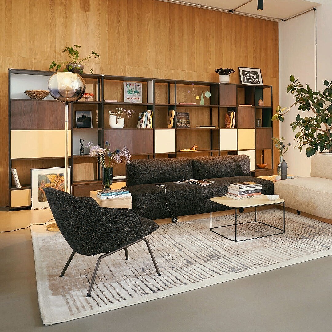 Modern lounge area with a white sofa, two black armchairs, a low coffee table on a patterned rug, a wooden shelving unit with books and plants, and large windows separating an adjacent seating area with pink chairs