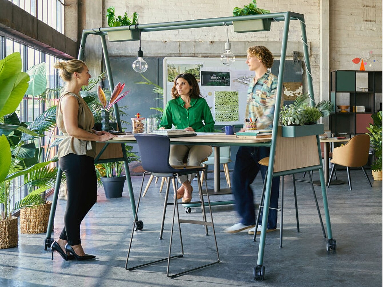 Drei Personen in einem modernen Büro mit vielen Pflanzen, die an einem hohen Tisch mit Metallgestell und Holzplatte arbeiten.