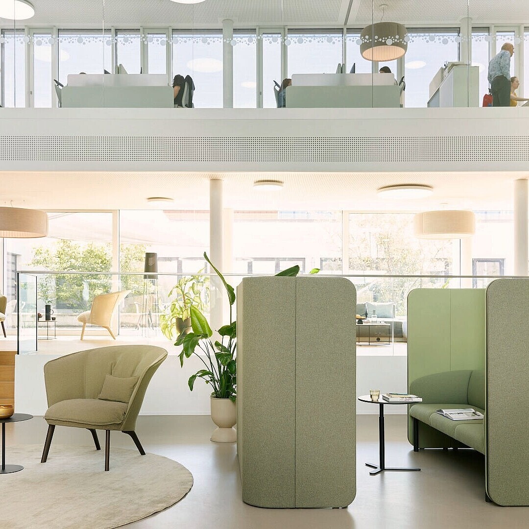Open-plan office space with seating areas separated by tall upholstered partitions and large windows providing natural light