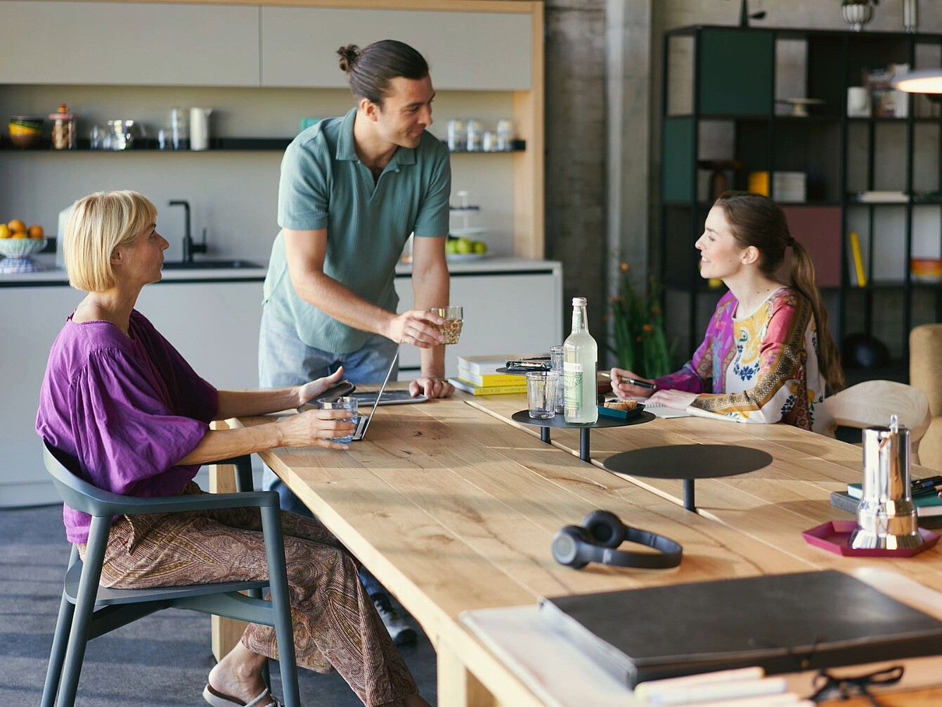 Three people in a kitchen around a wooden table with a laptop, glass bottles, and headphones