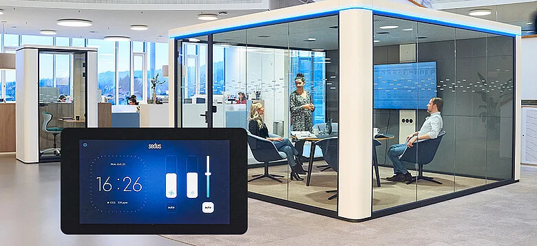 Modern office space with a glass-walled meeting room where three people are seated and one person is standing, with a digital touch display showing time and controls in the foreground.